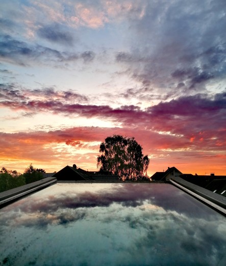 Farbenfroher Morgenhimmel über dem gekippten Dachfenster fotografiert. In der Mitte des Bildes leuchtet das  orange Morgenlicht durch eine Birke.
Der Himmel spiegelt sich im Fenster. Einiges Gewölk sorgt für kunstvolle Kontraste.